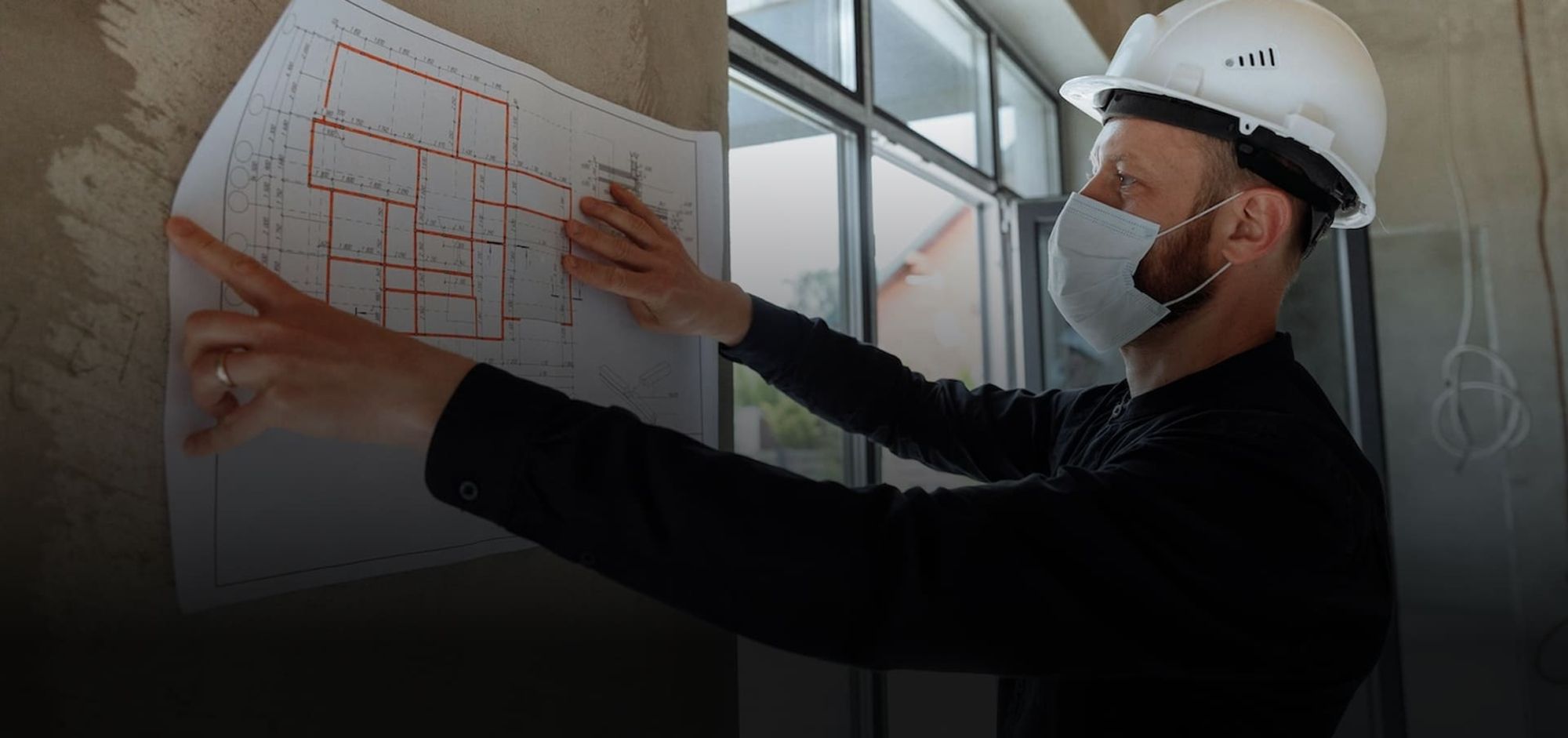 Ein Mann mit Schutzhelm und Maske studiert Baupläne an einer Wand auf einer Baustelle